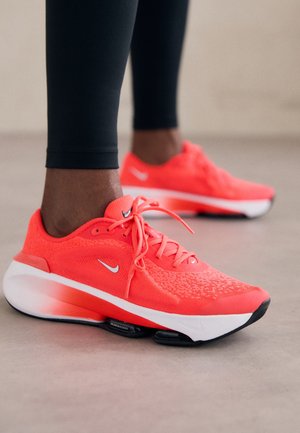 Zapatillas de running Nike de color naranja brillante usadas con mallas negras sobre un suelo interior liso, destacando el diseño de la suela acolchada.