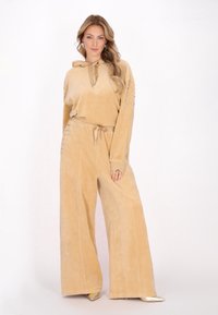 Tuta in velour beige con cappuccio, vita regolabile con cordoncino e pantaloni lunghi a gamba larga con dettagli a borchie lungo le maniche. Texture liscia.