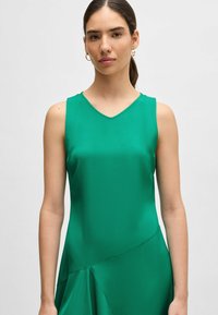 Zielona sukienka bez rękawów z dekoltem w kształcie litery V, gładką fakturą i asymetrycznym dołem. Materiał wydaje się lekki i elegancko się układa.