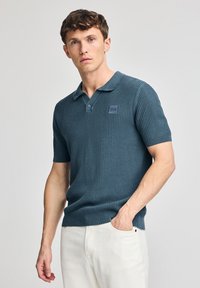 Camisa polo de manga corta en verde azulado oscuro, con textura acanalada, cuello clásico y un pequeño parche de logo en el pecho. Llevada con pantalones de color claro.