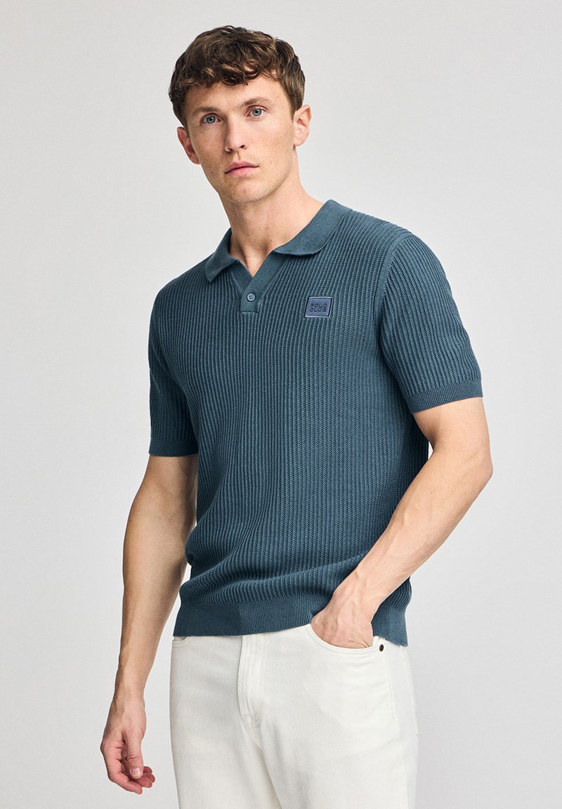 Camisa polo de manga corta en verde azulado oscuro, con textura acanalada, cuello clásico y un pequeño parche de logo en el pecho. Llevada con pantalones de color claro.