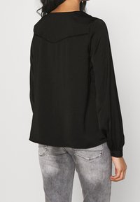 Blusa nera a maniche lunghe con colletto arrotondato e dettagli sul pattino, realizzata in tessuto liscio, abbinata a jeans grigi chiari.