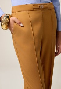 Pantalons marron ajustés avec une taille cintrée, de petites passants de ceinture, des poches avant et une texture lisse. Un bracelet doré orne le poignet.