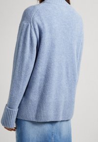 Pull en tricot bleu clair avec un ourlet et des poignets côtelés, doté d'une texture douce et d'un col montant. Porté par-dessus une jupe en denim.