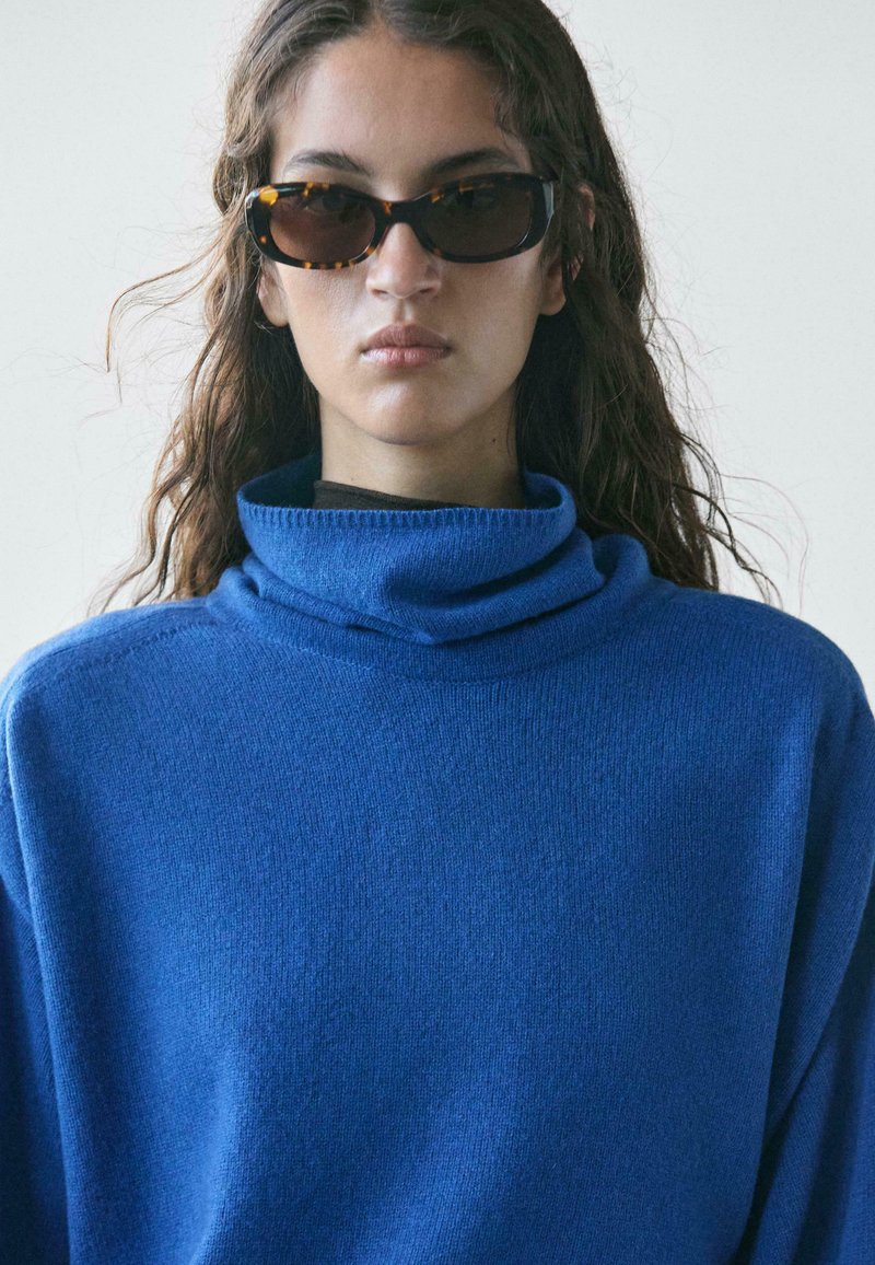 Giovane con capelli castani lunghi che indossa occhiali da sole tartarugati scuri e un maglione a collo alto blu brillante, su uno sfondo semplice.