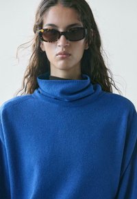 Junge Person mit langen braunen Haaren, die dunkle Schildpatt-Sonnenbrillen und einen leuchtend blauen Rollkragenpullover vor einem einfarbigen Hintergrund trägt.