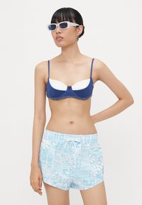 Modré a biele bikiny s pevným strihom a nastaviteľnými ramienkami, doplnené o vzorované svetlomodré plavky na šortky s kvetinovými motívmi.
