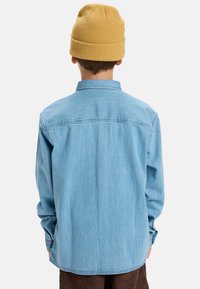 Persona vista desde atrás que lleva una camisa de mezclilla azul claro, pantalones marrones y un gorro de punto mostaza sobre un fondo liso.