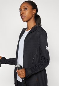 Black striped athletic jacket with a hood, zippered pockets, and thumbholes. Paired with a light grey shirt and holding trekking poles.