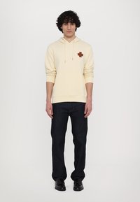 Cream-colored hoodie with a front pocket and a brown cross logo on the left chest, paired with dark blue jeans and black shoes.