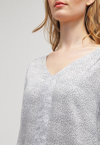 Lekka bluzka w białym kolorze z czarnym wzorem w kropki, z dekoltem w serek i subtelnym drapowaniem na ramionach. Gładka tekstura.