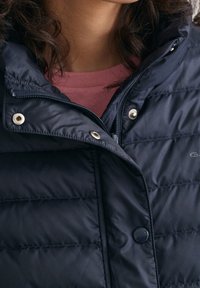 Schwarze gesteppte Jacke mit silbernen Druckknöpfen und Reißverschluss, über einem pinken Oberteil getragen, zeigt das untere Gesicht und den Hals einer Person.