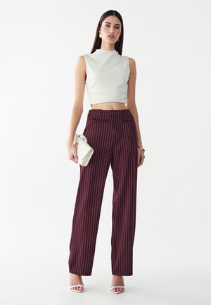 Donna che indossa un crop top bianco senza maniche, pantaloni larghi a righe bordeaux, sandali bianchi con tacco, che tiene una pochette intrecciata bianca.