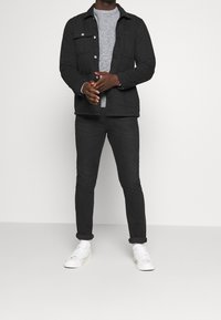 Schwarze knöpfbare Jacke mit vorderen Taschen, grauer strukturierter Pullover, schwarze Jeans und weiße Sneaker. Hochgekrempelte Jeans-Bündchen. Minimalistisches Design.