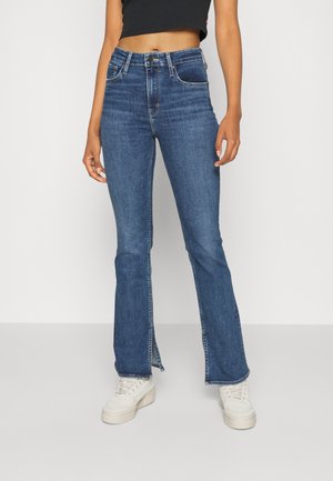 Kvinde iført højtaljede blå denimjeans med slidser foran ved kanten, kombineret med hvide sneakers og en sort cropped top.