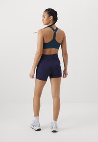 Teal sports-bh med justerbare stropper, kombinert med mørk lilla mønstrede shorts. Modellen har på seg hvite treningssko og ankelstrømper.
