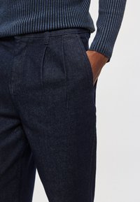 Tmavomodré riflové nohavice s hladkou textúrou, s plisami vpredu a bočnými vreckami. Nosené s rebrovaným námorníckym sveterom.