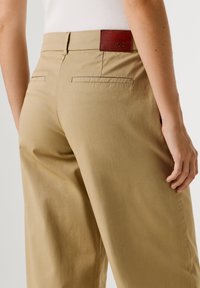 Μπεζ chinos με κομψό σχέδιο, διαθέτουν δερμάτινο ταμπελάκι στη μέση, δύο πίσω τσέπες και απαλό, υφασμάτινο ύφασμα με υφή.