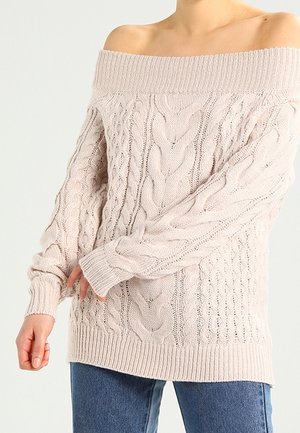 Femme portant un pull maille torsadée rose clair, à épaules dénudées, à manches longues, associé à un jean bleu.