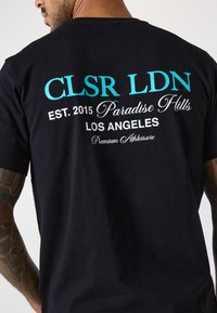 Svart bomull t-skjorte med hvit og teal tekst på ryggen: "CLSR LDN ETABLERT 2015 Paradise Hills LOS ANGELES Premium Athleisure."