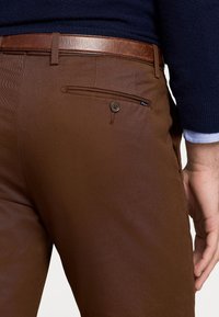 Bruna byxor i slätt tyg, med en knappfäst bakficka och ett brunt läderbälte. Designen är skräddarsydd med prydlig sömnad.