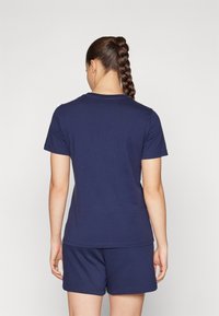 Kvinne med langt flettet hår, iført en enkel marineblå t-skjorte med korte ermer og matchende shorts, stående med ryggen mot kameraet foran en ensfarget bakgrunn.