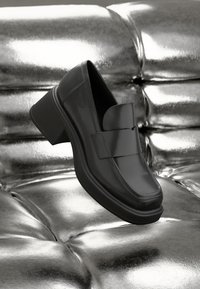 Black leather loafers with a wide strap and chunky heel, featuring a smooth finish and rounded toe, displayed on a metallic background.