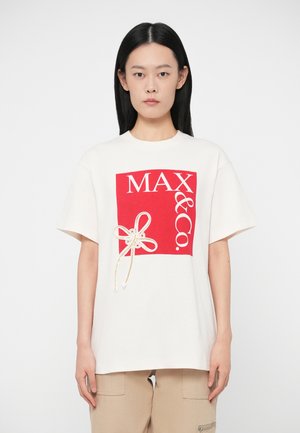 Frau mit glattem schwarzen Haar, die ein weißes T-Shirt mit einem roten "MAX & Co."-Quadratlogo und beigen Hosen vor einem weißen Hintergrund trägt.