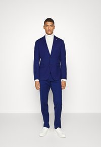 Blaues, maßgeschneidertes Anzug mit aufgeschlitztem Revers, einem Knopf und zwei Taschen. Kombiniert mit einem weißen Rollkragenpullover und weißen Sneakers.