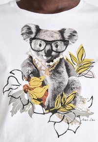 Balta marškinėliai su koalos grafika, dėvinčia akinius, papuošti perlų ir auksinės spalvos grandinėlės, taip pat geltonos ir pilkos spalvų gėlių siuvinėjimu.