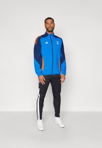 Chaqueta deportiva azul y marino con acentos naranjas y un logo, combinada con pantalones de chándal negros con franjas blancas y zapatillas deportivas blancas.