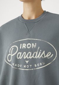 Grå hettegenser med ribbestrikket krage og mansjetter. Har et slitt hvitt grafisk trykk med teksten "IRON Paradise MADE NOT BORN" i et ovalt design.
