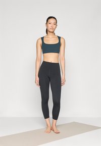 Conjunto atlético que incluye un top corto de un verde azulado oscuro con tirantes anchos y leggings negros de largo completo. Textura suave, diseño ajustado.