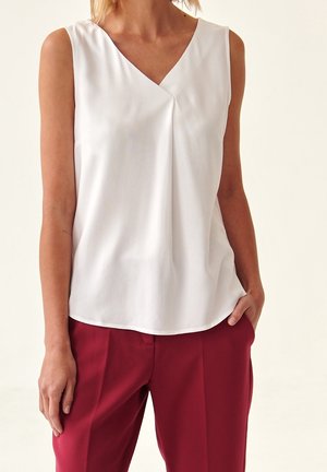 Biała bluzka bez rękawów z dekoltem w serek i plisą z przodu, noszona z wysokim stanem bordowymi spodniami, jedna ręka w kieszeni.