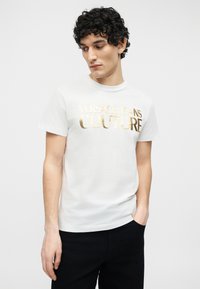 Νέος άνδρας με σγουρά μαλλιά που φοράει λευκό T-shirt με χρυσό λογότυπο "Versace Jeans Couture", μαύρο παντελόνι, και έχει το ένα χέρι στην τσέπη.