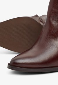 Bottine en cuir marron avec un bout pointu, texture lisse et semelle en caoutchouc plate dotée de détails rainurés en dessous pour une meilleure adhérence.