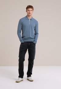 Een jonge man met krullend haar staat zelfverzekerd, gekleed in een lichtblauwe trui, donkere jeans en sneakers, tegen een neutrale achtergrond.