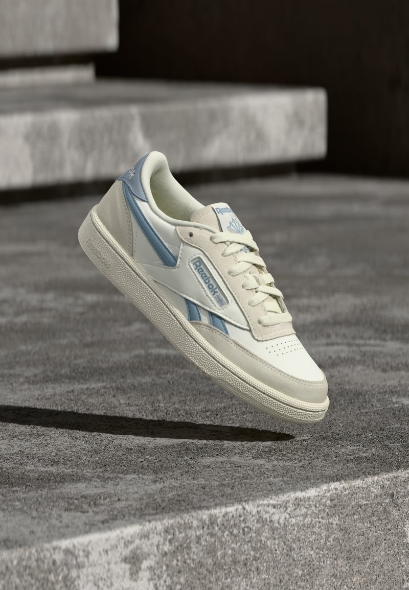 Bijela i svijetloplava Reebok tenisica suspendirana iznad betonskog stepenika s pozadinom od teksturirane sive podloge.