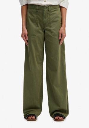 Donna che indossa pantaloni larghi verde oliva con tasche frontali applicate e sandali neri, in piedi dritta su uno sfondo bianco.