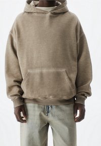 Sudadera de forro polar marrón con un bolsillo frontal, capucha con cordón y puños de canalé, combinada con unos pantalones vaqueros anchos de lavado claro. Diseño minimalista.