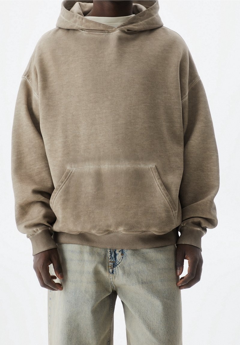 Sudadera de forro polar marrón con un bolsillo frontal, capucha con cordón y puños de canalé, combinada con unos pantalones vaqueros anchos de lavado claro. Diseño minimalista.