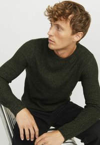Dunkelgrüner Strickpullover mit geripptem Muster und Rundhalsausschnitt, kombiniert mit schwarzen Hosen. Das Design verfügt über lange Ärmel und eine figurbetonte Silhouette.