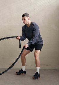 Schwarze Trainingsseile werden von einer Person in einem grauen Langarmoberteil und schwarzen Shorts verwendet, die auf einem hellgrauen Boden steht.