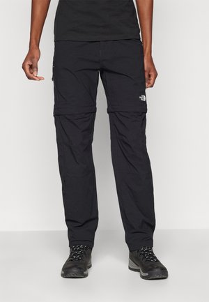 Person wearing black convertible hiking pants and black hiking boots, standing against a plain light background.