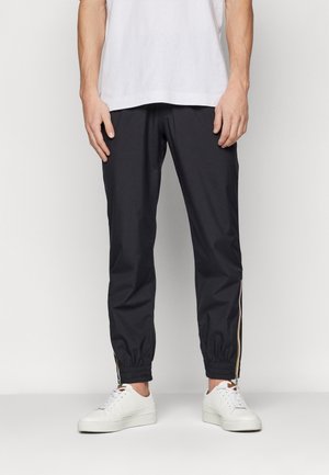Mand iført sorte joggerbukser med elastiske manchetter og hvide sneakers, stående mod en ensfarvet lys baggrund.