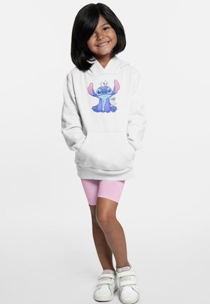 Vit hoodie med en grafik av en blå karaktär med rosa öron på framsidan. Bärs över rosa shorts med vita skor. Barnet står och ler.