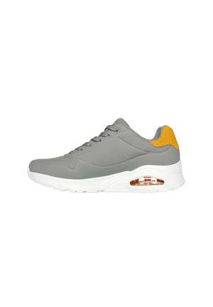 Zapatilla deportiva gris con material suave, suela blanca y acento amarillo en el talón. Cuenta con detalles perforados y cordones.