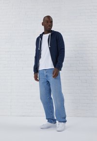 Felpa con zip blu navy e cappuccio, con lacci bianchi, t-shirt bianca, jeans azzurri chiari e sneakers bianche, su uno sfondo di mattoni bianchi.