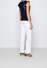 Námořnicky modrý bezrukávový top s volánky a bílým lemem, doplněný o bílé široké kalhoty a černé sandály na podpatku, zobrazený zezadu.