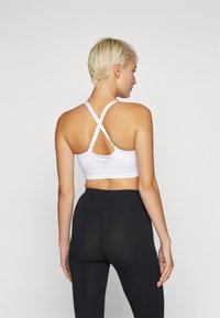 Hvit sports-BH med kryssede stropper, med en jevn tekstur og minimalistisk design. Brukt med svarte høyhælte leggings.